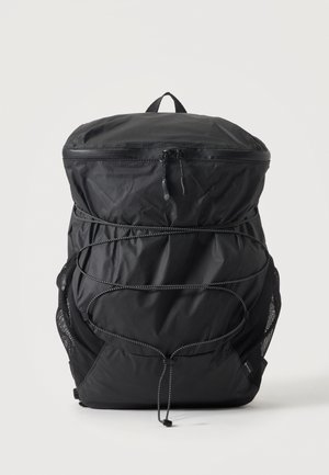 Sac à dos léger noir avec fermeture éclair sur le dessus, cordons élastiques réglables à l'avant, poches latérales en maille et poignée de transport supérieure sur fond blanc.