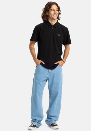 Polo noir avec un col et un petit logo sur la poitrine, associé à un jean en denim bleu clair ample et des baskets noir et blanc.
