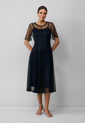 Robe bleu marine avec des manches courtes et transparentes, dotée d'un corsage ajusté qui se prolonge en une jupe plissée fluide. Sandales à talons argentées.