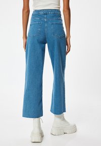 Jeans de pierna ancha en denim azul medio con una cintura alta, bolsillos traseros y detalle de dobladillo sin terminar, combinados con botas blancas gruesas.