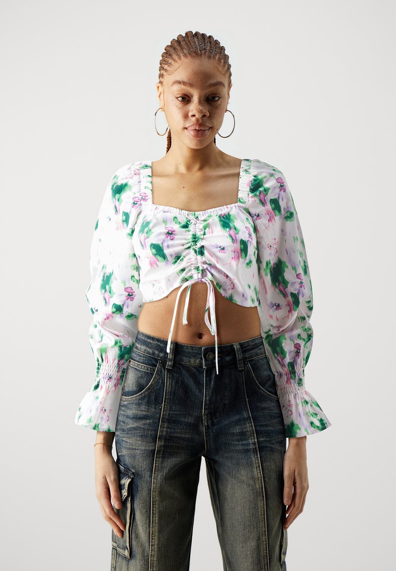 Kvetinový crop top so štvorcovým výstrihom, vypchatými rukávmi, zelenými a ružovými vzormi, nazberanými detailmi a viazaním vpredu. Nosený s tmavými džínami.