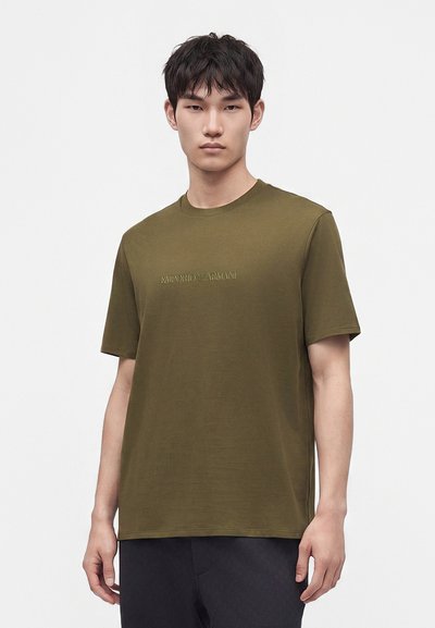 Млад мъж, облечен в маслиненозелена тениска Emporio Armani с къси ръкави и черен панталон, стоящ срещу обикновен бял фон.