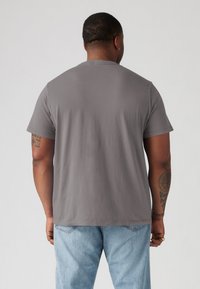 Grijze katoenen T-shirt met korte mouwen, ronde hals en een losse pasvorm. Zichtbare tatoeages op de arm en lichtblauwe spijkerbroek.