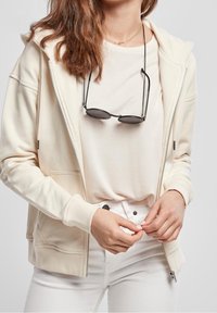 Krémová mikina na zip z měkké tkaniny, spárovaná s světlým béžovým tričkem, nošena s bílými džíny. Sluneční brýle visí na krku na řetízku.