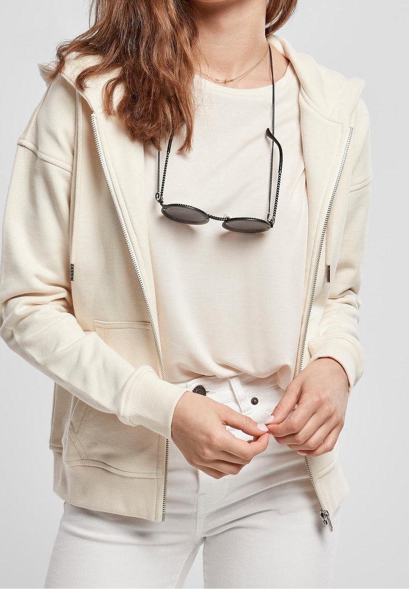 Krémová mikina na zip z měkké tkaniny, spárovaná s světlým béžovým tričkem, nošena s bílými džíny. Sluneční brýle visí na krku na řetízku.
