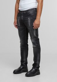 Mann iført svarte slitte jeans med hvite malingssprut, svarte loafers og en hvit ermeløs skjorte mot en ensfarget bakgrunn.