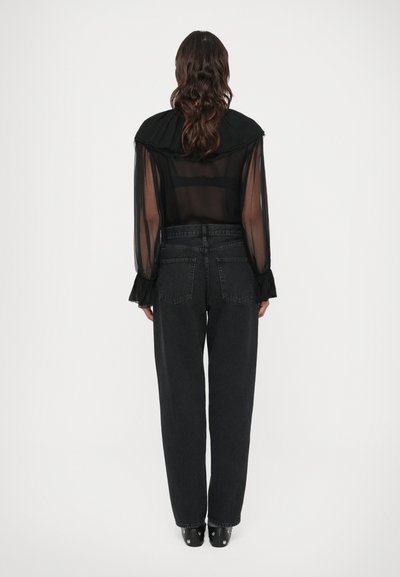 Blusa preta transparente com punhos franzidos e gola alta, combinada com jeans pretos de cintura alta e saltos grossos pretos.