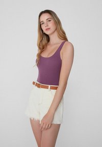 Mujer con cabello rubio largo vistiendo una camiseta de tirantes morada y pantalones cortos de mezclilla blancos de talle alto con un cinturón marrón, de pie contra un fondo liso.