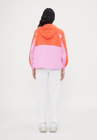 Color-blocked hooded jacket features orange and pink panels, lightweight fabric, relaxed fit, and elastic cuffs. Worn with white pants.