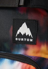 Burton branded backpack detail featuring a black label with a white mountain logo on a colorful, textured background with gradient shades.