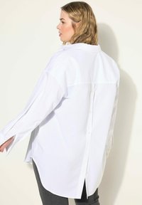 Hvid skjorte med knapper og slids bagpå, lange ærmer og buet kant. Fremstillet af glat stof med diskrete knapper langs ryggen.