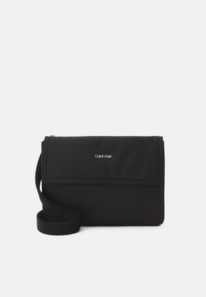 Zwarte Calvin Klein crossbodytas met klepafsluiting en verstelbare schouderriem op een effen lichte achtergrond.