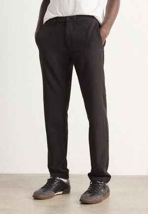 Homme portant un pantalon noir slim et des baskets noires et grises, debout les mains dans les poches devant un fond uni.
