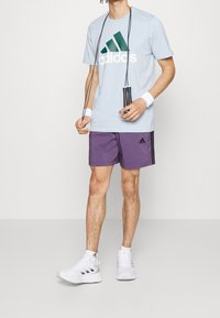 Světle modré sportovní tričko s zeleným a bílým logem Adidas, doplněné fialovými šortkami s černými pruhy a bílými sportovními botami.