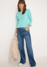 Camiseta de manga larga color verde menta, jeans de pierna ancha de lavado medio, sosteniendo una chaqueta color crema y vistiendo zapatillas bajas blancas.