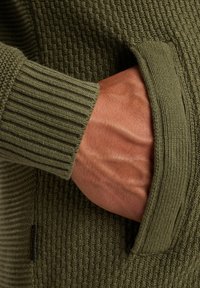 Hand teilweise in der vorderen Tasche eines olivgrünen strukturierten Strickpullis mit gerippten Bündchen und Taschenrändern.