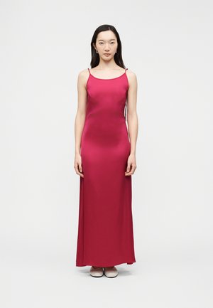 Woman with long dark hair wearing a sleeveless, floor-length bright pink slip dress and light-colored flat shoes, standing against a plain background.