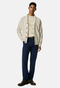 Beige Strickjacke mit Schalkragen, Knopfverschluss, über einem cremefarbenen gestreiften Hemd getragen, kombiniert mit navy Hosen und braunen Schuhen.
