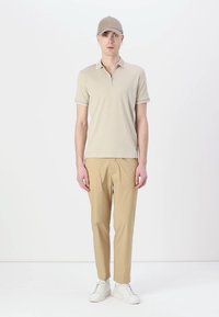 Beige polo skjorte med hvit kant, sammen med beige bukser. Hvite joggesko. Modellen står mot en enkel hvit bakgrunn.