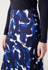 Personne portant une jupe à motif abstrait bleu, noir et blanc avec un haut noir à manches longues, vue de la taille jusqu'au milieu de la cuisse.