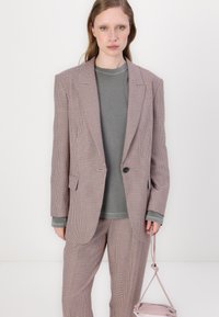 Blazer xadrez em tons de rosa e cinza com um design estruturado, combinado com um sweatshirt cinza e calças a condizer. Acessório de mala leve.