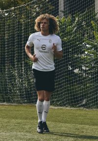 Maglia da calcio bianca con emblema e logo, pantaloni corti neri, calze bianche e scarpe nere. Giocatore con capelli ricci sta correndo su un campo in erba verde.