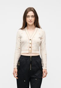 Νεαρή γυναίκα με μακριά καστανά μαλλιά, φοράει cropped μπλε ριγέ ζακέτα με χρυσά κουμπιά και μαύρο παντελόνι με λεπτομέρειες από χρυστό φερμουάρ.