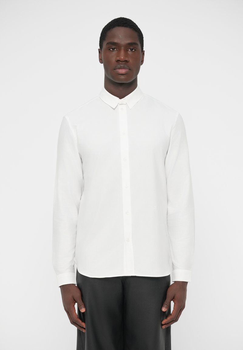 Chemise blanche à manches longues avec boutons, en tissu lisse, dotée d'un col classique et d'un ourlet droit, associée à un pantalon noir.