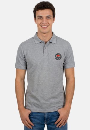 Jeune homme portant un polo gris avec un petit logo circulaire sur la poitrine gauche et un jean bleu, souriant avec les mains près des poches.