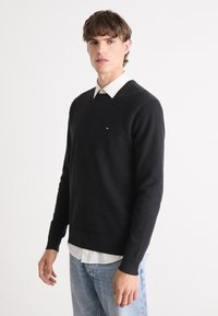 Zwarte gebreide trui met een ronde hals, voorzien van ribbelmanchetten en zoom. gedragen over een witte blouse met kraag. Klein logo op de borst.