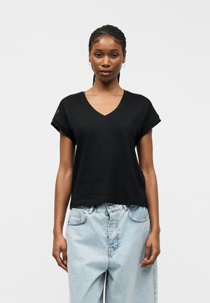 Ung kvinde med fletninger iført en sort V-hals T-shirt og lyseblå højttaljede jeans, stående foran en ensfarvet baggrund.