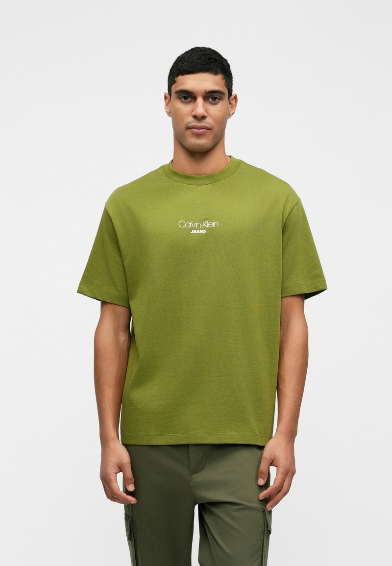 Zelena pamučna majica s okruglim izrezom, kratkim rukavima i bijelim logom "Calvin Klein JEANS" na prednjem dijelu. Ima opušteni kroj.