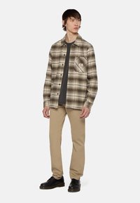 Geruite flanellen shirt in beige en bruine tinten, voorzien van een borstzak en zwarte knopen. Draagt over een donker T-shirt met tan kleurige broek en zwarte schoenen.