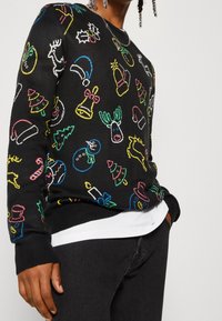 Eine Person trägt einen schwarzen Pullover mit bunten gestickten Weihnachtsmotiven, darunter Rentiere, Schneemänner, Glocken, Bäume und Zuckerstangen.