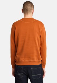 Felpa arancione a girocollo con polsini e orlo a coste, realizzata in un tessuto morbido. Indossata con jeans in denim scuro, con tasche sul retro.