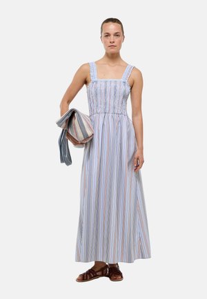 Femme portant une robe longue sans manches rayée verticalement bleu et blanc, tenant dans un bras un textile plié assorti rayé.