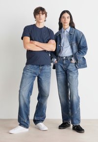 To modeller står side om side. Mannen har på seg en marineblå t-skjorte og lyseblå jeans; kvinnen har på seg en dongerijakke, lyseblå skjorte og jeans.