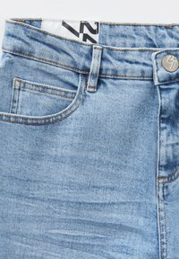 Jeans in denim azzurro chiaro con vita alta, tasca frontale, cuciture arancioni e bottone in metallo. L'etichetta presenta un design in bianco e nero.