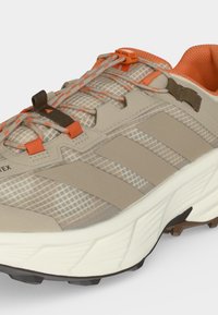 Chaussure de trail beige et orange avec tige en mesh à motifs, système de laçage rapide, col rembourré et semelle blanche épaisse avec une semelle extérieure robuste.