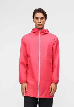 Άνδρας που φοράει μακρύ ροζ αδιάβροχο μπουφάν με κουκούλα, με μπροστινό φερμουάρ και φερμουάρ σε πλαϊνές τσέπες, στέκεται μπροστά σε μονόχρωμο γκρι φόντο.