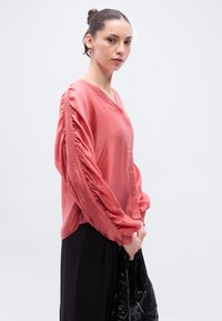 Κοραλί πουκάμισο με κουμπιά και βολάν στα μανίκια, κατασκευασμένο από ελαφρύ ύφασμα. Το μοντέλο το συνδύασε με μαύρο παντελόνι και μαύρη τσάντα με υφή.