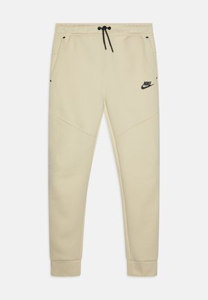 Beige Nike-housut elastisella vyötäröllä ja kiristysnauhalla, joissa on sivutaskut, musta logo ja tekstuuripaneelikorostukset.