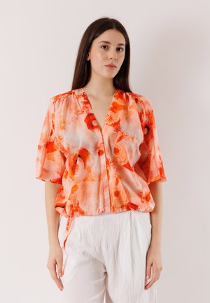 Donna con capelli lunghi e scuri indossa una blusa arancione floreale a maniche corte e pantaloni bianchi con trama, in piedi contro uno sfondo chiaro e semplice.