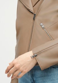 Μπεζ δερμάτινο μπουφάν με ασημένια φερμουάρ στο μπροστινό μέρος, στην τσέπη και στην μανσέτα, φορεμένο πάνω από μπλε τζιν παντελόνι, εμφανιζόμενο στο αριστερό χέρι και στο μπροστινό μέρος του σώματος.