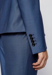 Costume bleu sur mesure avec un tissu texturé. Boutons noirs sur le poignet, finition lisse, coupe ajustée, gros plan sur la manche de la veste.