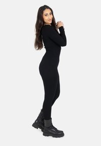 Zwart geribbeld jumpsuit met een aansluitend ontwerp, gecombineerd met zwarte veterschoenen met combat-stijl. Het materiaal lijkt rekbaar en gestructureerd.