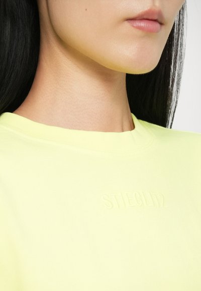 Yellow cotton t-shirt with a round neckline. Features embossed logo "STIEGLITZ" on the front. Smooth texture and fitted design.