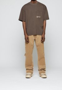 T-shirt en coton marron avec une poche poitrine et un détail texte, associé à un jean en denim beige avec des bords effilochés et une coupe ample.