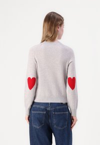 Maglione knit grigio chiaro con maniche lunghe e motivi a cuore rossi sui gomiti. Collo e orlo a costine. Abbinato a jeans blu.