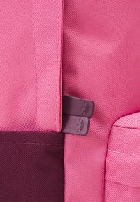 Vaaleanpunainen kangaskassi, jossa on teksturoitu pinta, tumma vaaleanpunainen korostus ja kaksi purppuranväristä vetoketjua sivuilla. Yksinkertainen ja toimiva muotoilu.